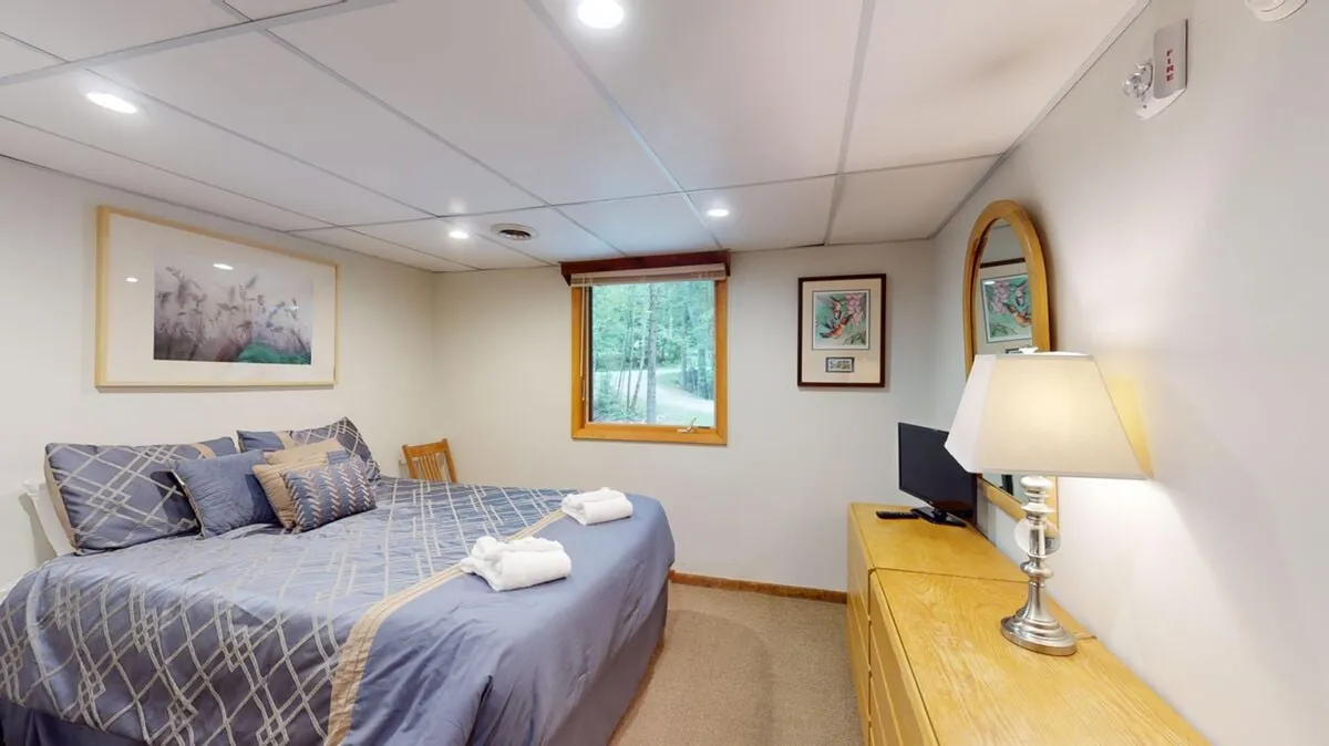 A tranquil bedroom with a plush bed adorned with decorative pillows, a window letting natural light stream in, artwork on the wall, and a dresser with a lamp. The overall mood is calm and serene, suggesting a restful environment perfect for unwinding.