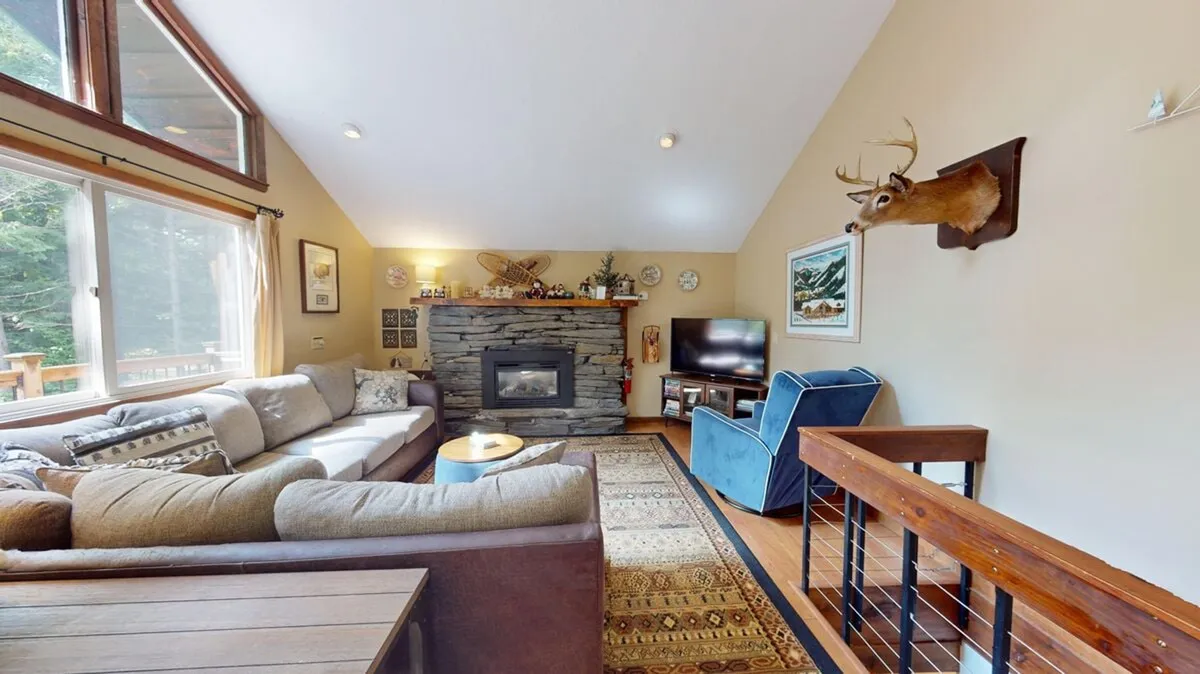 The image features a spacious and inviting living room with a large sectional sofa arranged around a striking stone fireplace, a mounted deer head above the mantle, and rustic wooden railings. The warm lighting and cozy furnishings set a welcoming, mountain-chalet ambiance perfect for gatherings and relaxation.