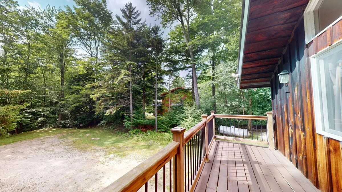 A spacious wooden deck is nestled among dense trees, with a gravel driveway and a glimpse of a neighboring cabin. The deck is set up for relaxation, outdoor meals, and gatherings, inviting guests to unwind or prepare for adventure. The surrounding landscape immerses viewers in the Vermont mountain setting.