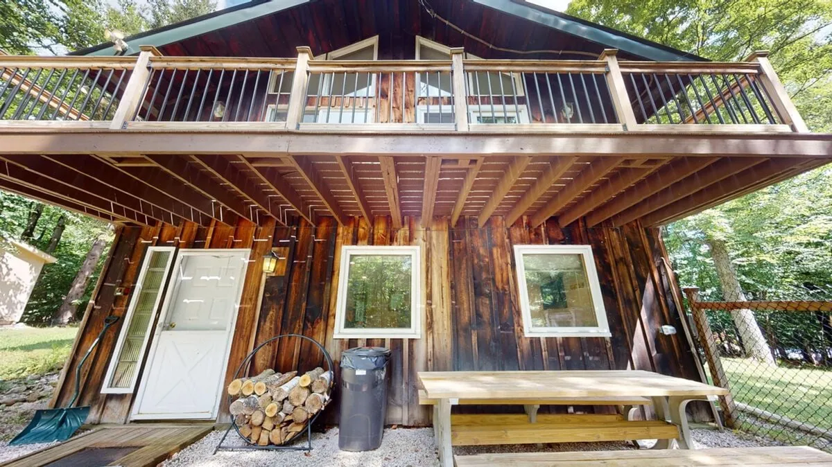 This exterior image showcases the chalet's welcoming wooden balcony, visible front door, a stack of firewood, and a picnic table, all backed by a tranquil forest setting. The scene conveys an inviting space perfect for relaxing outdoors after a day on the slopes.