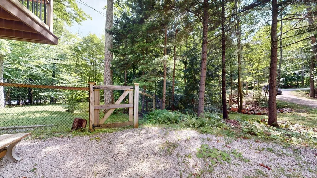 This image showcases a wooden gate that forms part of a sturdy fenced area, with a gravel path leading inside. Tall trees and lush greenery surround the space, creating a tranquil atmosphere. The fence is clearly visible, emphasizing the secure outdoor area ideal for pets to roam, while the natural landscape reinforces the mountain setting.