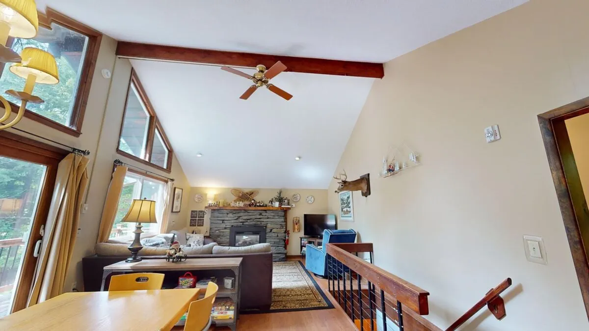 This image features a living area with a fireplace, comfortable sofa, dining table, ceiling fan, and expansive windows. The layout encourages both relaxation and group interactions, with the fireplace anchoring the space for winter gatherings and the windows suggesting a connection to the outdoors.