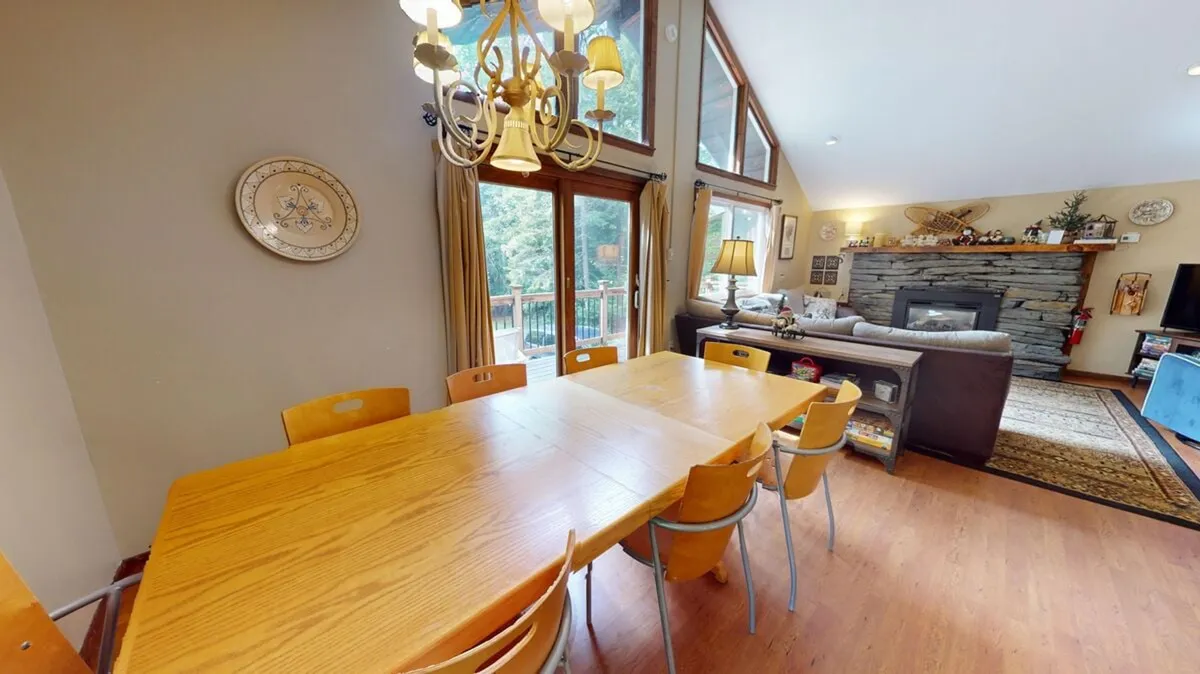 This image features a large dining table set beneath a chandelier, with a backdrop of large windows and a prominent stone fireplace. The space is ideal for group meals, game nights, and holiday celebrations, with ample natural light and a welcoming, communal atmosphere.