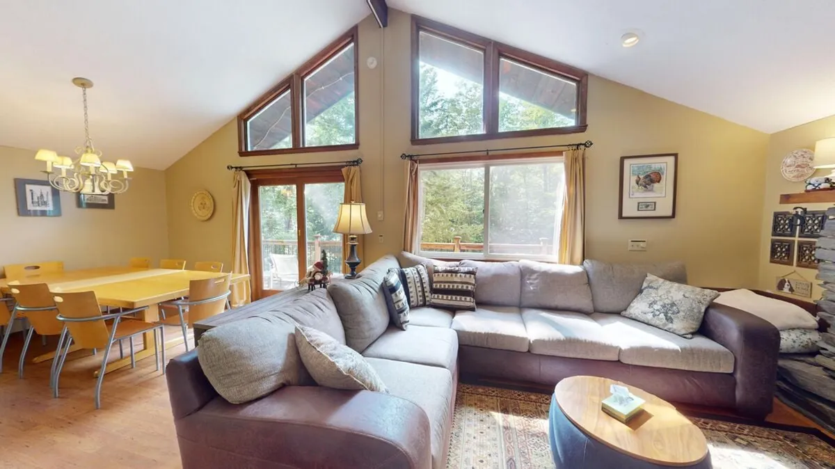 This image showcases a spacious open living area featuring a large sectional sofa, a dining table, ample natural light streaming through the windows, and decorative shelving. The space is clearly designed for group activities such as family gatherings, socializing, and relaxation, with the living area and dining space seamlessly connected.