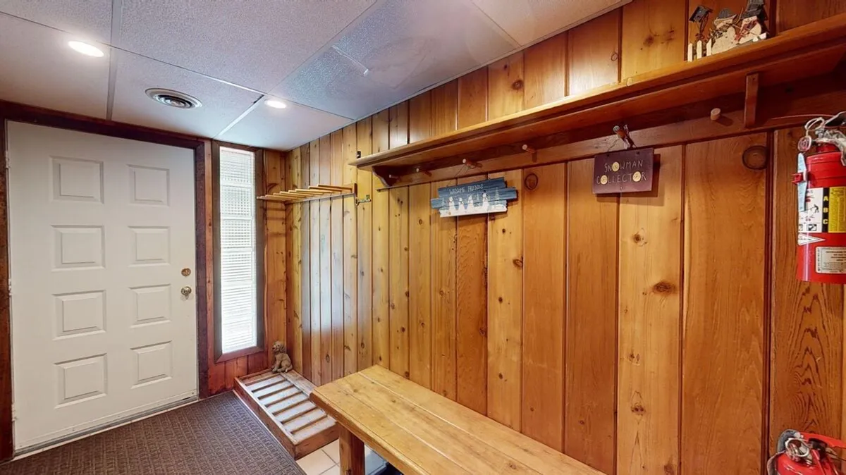 This interior image captures the entryway, featuring a wooden bench, shelves, an entrance door, and a welcoming decorative sign. It evokes the moment of returning home after a day out, with space for gear and a cozy atmosphere.