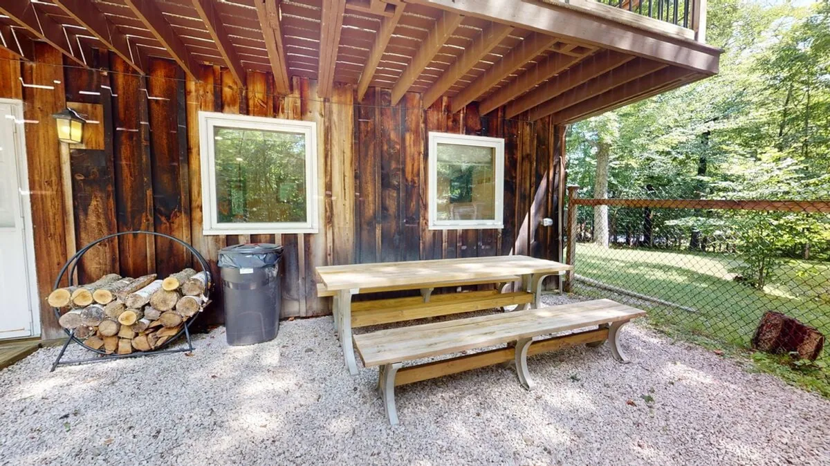 A wooden picnic table sits beside a stack of firewood, adjacent to the house’s exterior with large windows letting in light. The setting suggests a cozy spot for outdoor meals, family gatherings, or simply enjoying the fresh air with pets nearby. The blend of natural and homey elements brings outdoor comfort to life.